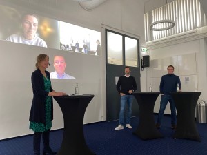 Eine Frau und zwei Männer mit Mikrofonen stehen und sprechen zu einem Publikum. Zwei weitere Personen sind per Videocall zugeschaltet.